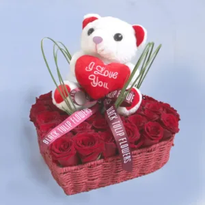 The image shows a heart-shaped basket filled with red roses. A white teddy bear is sitting on top of the roses, holding a heart that says "I Love You.