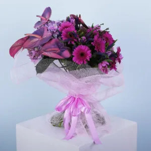 purple flowers is arranged in a rounded, full shape and wrapped in light purple mesh fabric. The bouquet features purple anthuriums, purple gerbera daisies, and smaller purple flowers with a feathery texture