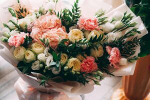 A close-up shot of a lush bouquet of flowers being held by someone. The bouquet features a mix of light pink and white carnations, creamy yellow roses, and various green foliage.