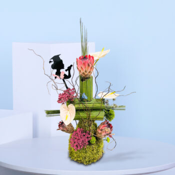 Graduation-themed flower arrangement with proteas, pink anthuriums, moss base, and a silhouette of a graduate holding a diploma, styled with natural twigs and butterflies.