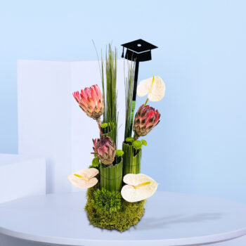 Graduation-themed floral arrangement with pink proteas, white anthuriums, tall green foliage, moss base, and a black graduation cap topper.