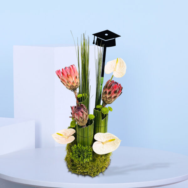 Graduation-themed floral arrangement with pink proteas, white anthuriums, tall green foliage, moss base, and a black graduation cap topper.