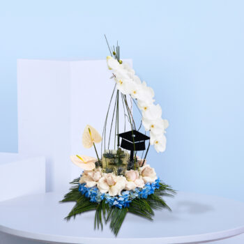 Graduation-themed floral arrangement with white orchids, blush roses, pink anthuriums, blue hydrangeas, and a black graduation cap topper.