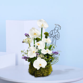 Graduation-themed floral arrangement with white orchids, greenery, and purple accents in a moss base, featuring a line art of a graduate in the background.
